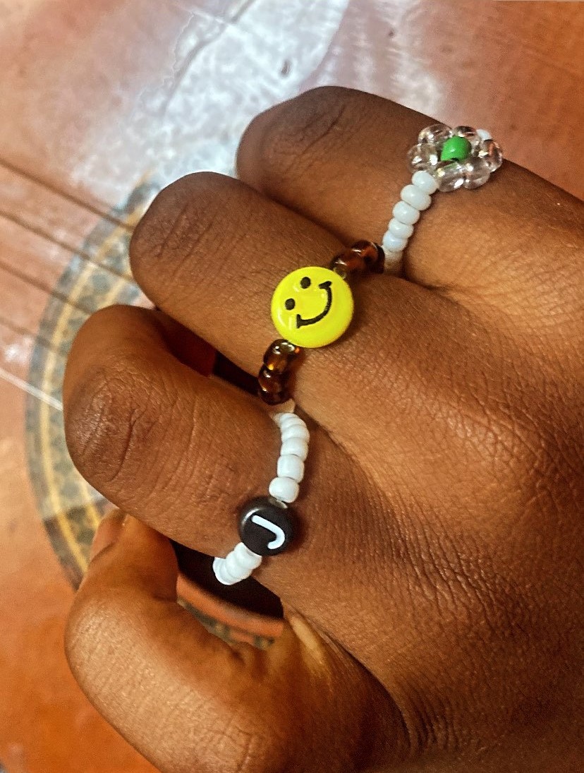 Hand wearing three beaded bracelets on the fingers, one with a smiley face bead and the other with a letter ‘J’ charm, and the last one looks like a ring with a green stone on the pinky finger